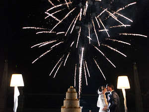 alt=The bride and groom cut the cake with fireworks in the background at Villa Osconi Bertani, Lake Garda